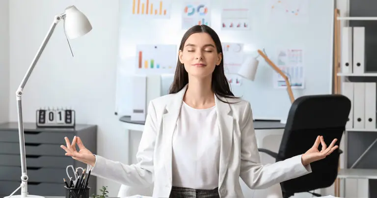 A professional woman in a white blazer sits peacefully in a modern office environment with her eyes closed, practicing mindfulness meditation. Her hands rest on her knees, palms up, with her fingers forming a meditation pose. The workspace behind her features a desk lamp, office supplies, charts on the wall, and a clean, organized atmosphere, conveying a sense of productivity and calm all to represent employee wellness trends for 2025.