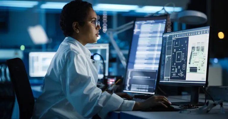 Image of a woman with short hair wearing a white long sleeve shirt sitting in front of two computer monitors displaying many lines of data to represent data science and data scientists