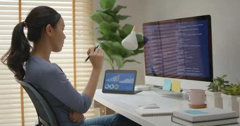 A woman with long dark hair tied back in a ponytail sits at a modern white desk, looking intently at a large desktop monitor displaying lines of code. She holds a pen in her right hand, appearing to be thinking or analyzing the code. The desk setup includes a laptop showing charts and graphs, a notebook, a white keyboard and mouse, a pink mug on a coaster, and small potted plants. A large green plant is visible in the background, and natural light streams in through blinds, creating a bright and focused workspace.