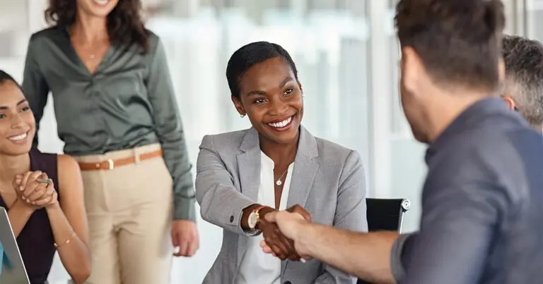 A group of diverse professionals in a modern office setting are engaged in a business meeting. A woman in a gray blazer and white blouse is smiling and shaking hands with a man in a navy blue shirt, indicating a successful interaction. Two other colleagues, a woman in a dark sleeveless top and another in a green blouse and beige pants, are observing the handshake with positive expressions. The setting is well-lit with natural light and features a professional atmosphere.