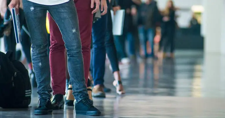 Photo of the bottom half of a group of people seemingly waiting in line. The individual in front is wearing blue jeans and black sneakers while the person behind them wears red pants and black shoes.