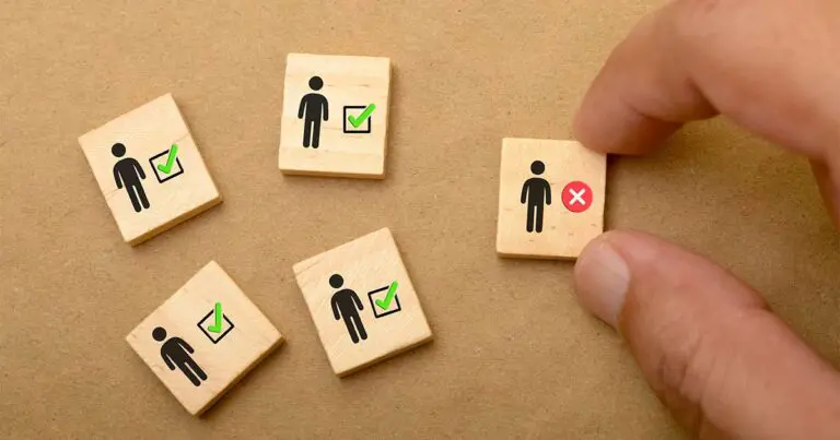 A hand picks up a wooden block with a person icon and a red "X," while four other blocks with person icons and green checkmarks remain on a brown surface. The image symbolizes candidate selection, hiring decisions, or applicant tracking system filtering, highlighting the process of choosing the right candidate while rejecting others.