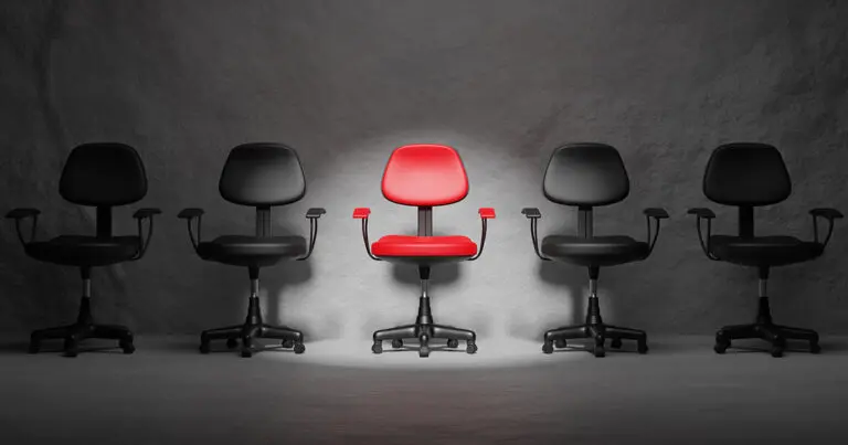 A single red office chair sits in the center of a row of four black chairs, all positioned against a dark, textured background. A spotlight highlights the red chair, emphasizing its uniqueness. The image symbolizes standing out from the competition, the hiring process, or the challenge of finding the right candidate for a role without falling victim to outdated hiring myths.