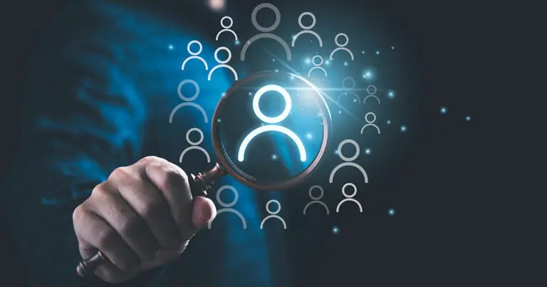 A hand holding a magnifying glass focuses on an illuminated person icon, surrounded by smaller, transparent person icons on a dark background. The image symbolizes talent acquisition, recruitment, or searching for the right candidate. Bright blue tones and glowing elements convey a modern, digital theme.