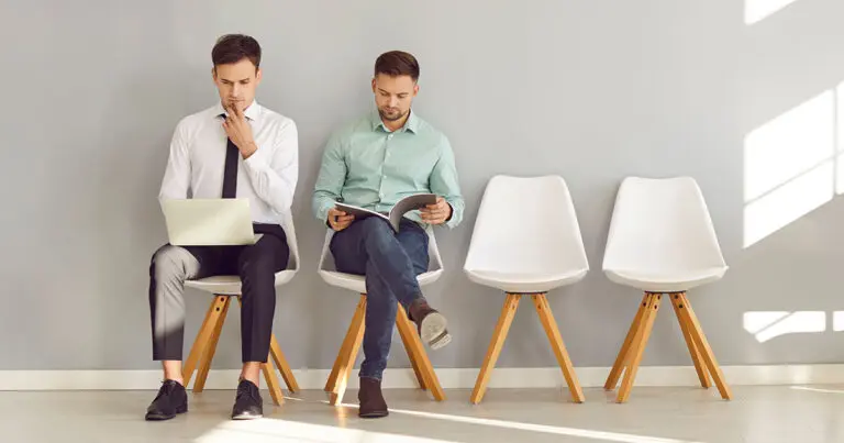 Two men sit side by side in a bright, minimalist waiting room with four white chairs lined against a light gray wall. One man, dressed in a formal white shirt and black tie, works intently on a laptop, while the other, in a casual mint-green shirt and jeans, reads a magazine. The contrasting outfits and activities suggest different approaches or styles, often seen in job interviews or hiring processes. Empty chairs beside them highlight the simplicity and focus of the scene.