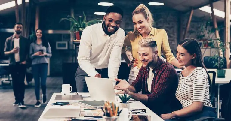 A diverse group of young professionals gathers around a laptop in a modern, open office space, smiling and collaborating on a project. Two team members stand in the background, holding coffee cups and observing the scene. The environment features warm lighting, plants, and exposed brick walls, creating a welcoming and creative atmosphere.