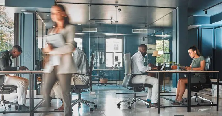 A busy office scene in a brightly lit room with a blue accent wall and 2 workers sitting at a table with one blurred worker walking past the camera to indicate movement and the fast pace of the office.