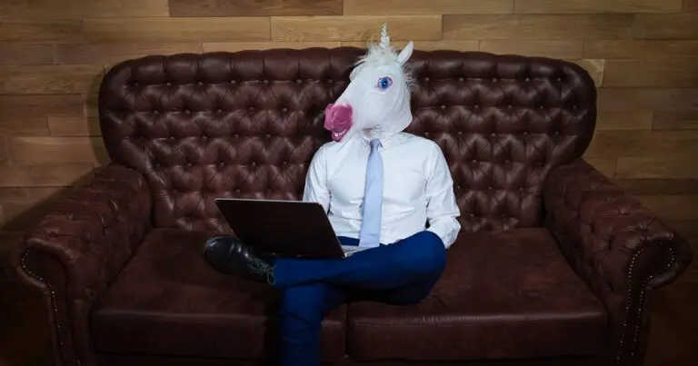 A person wearing a white unicorn mask, dressed in business attire with a white shirt, light blue tie, and navy blue pants, sits cross-legged on a brown leather couch while using a laptop. The setting features a wood-paneled wall, giving a professional yet whimsical feel. The image humorously represents the idea of searching for a "unicorn" candidate or a perfect candidate in hiring or the unrealistic expectations of finding the perfect candidate.