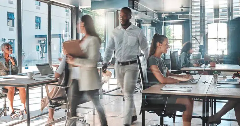 A modern office setting with large windows letting in natural light. Several professionals are actively working at desks, while others move around the space, creating a sense of energy and collaboration. One person types on a laptop, while another carries a folder, and others engage in discussions. The bright and open atmosphere reflects productivity and teamwork.