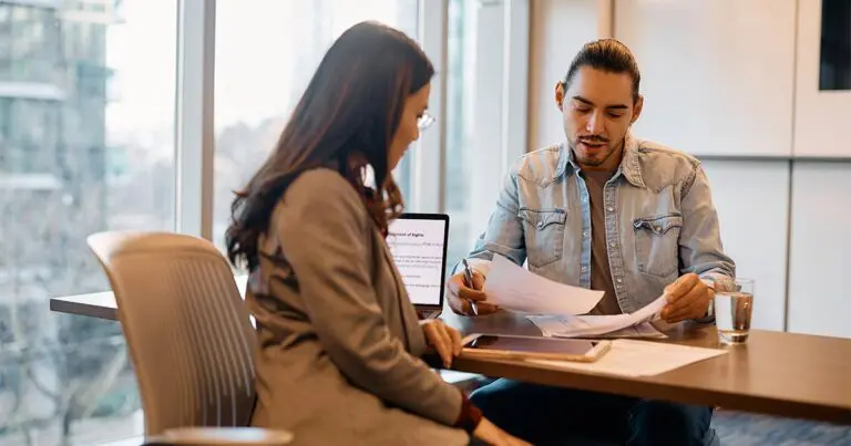 A professional man and woman sit across from each other at a table in a modern office with large windows. The man, wearing a denim jacket, reviews documents while speaking, while the woman listens attentively. A laptop with visible text is open on the table, alongside papers and glasses of water. The setting suggests a job interview, contract discussion, or business meeting.