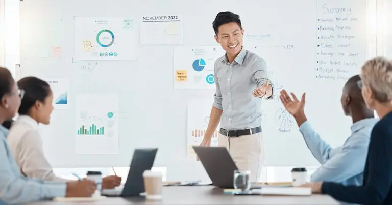 A young professional leads a presentation in a modern office, smiling and engaging with colleagues seated at a conference table. A whiteboard behind him is filled with charts, graphs, and strategy notes. Team members, diverse in age and background, participate in the discussion, reflecting a collaborative and data-driven work environment focused on marketing, analytics, or business strategy.