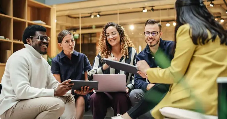 A diverse group of professionals sits in a casual, modern office space, engaged in a collaborative discussion. Some hold tablets and notebooks, while one person works on a laptop. The group is smiling and actively exchanging ideas, symbolizing teamwork, innovation, and a positive workplace culture. The setting features warm lighting and an open, contemporary design.