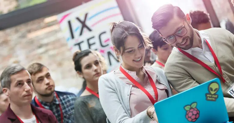 Image of a group of young professionals at a tech conference, wearing red lanyards. Two individuals in the foreground are smiling and engaging with a laptop covered in stickers. The background features other attendees and a colorful sign reading 'IX TECH.' The setting reflects collaboration, innovation, and networking in the tech industry.