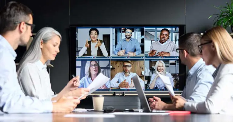 A group of four professionals sits in a modern conference room, engaging in a hybrid meeting with six remote colleagues displayed on a large screen. The remote participants, appearing in video call windows, actively gesture and speak. The in-person attendees review documents and take notes, symbolizing a blended workforce and the integration of remote and on-site collaboration.