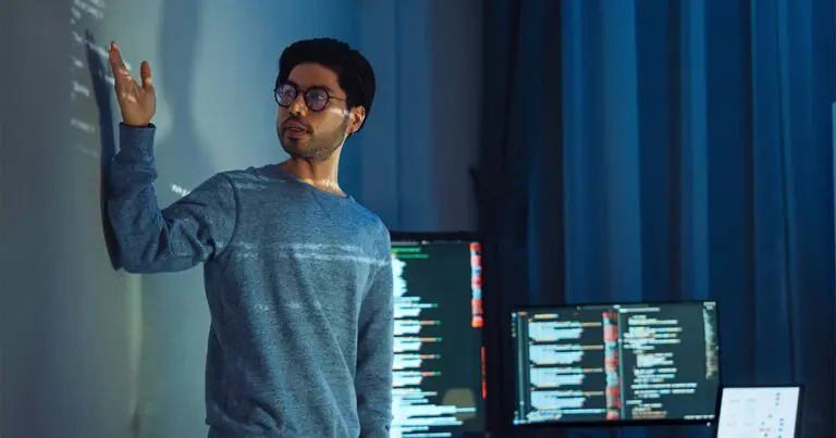 A man with short dark hair and glasses wearing a blue long-sleeve shirt stands on a stage with computer monitors displaying code behind him and what looks like computer code projected onto him and a large screen that he gestures toward to represent the future of AI jobs and the decline of prompt engineering.