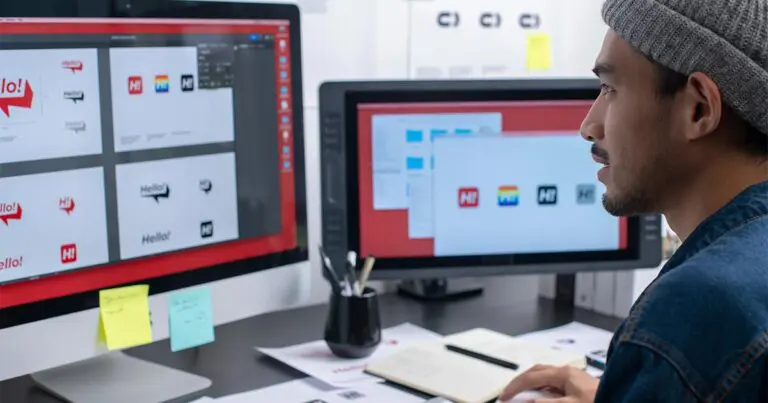 Two computer monitors with various logo designs and color swatches sit upon a black desk that is covered in papers with what look to be fonts and designs as a young man sits at the desk reviewing the designs and papers to represent the different types of graphic design.