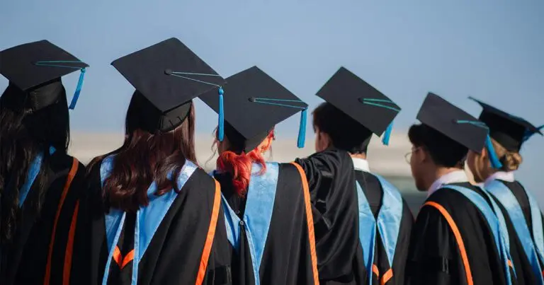 6 college graduates standing shoulder to shoulder in the traditional cap and gown with blue sashes facing away from the camera to symbolize looking forward into the future