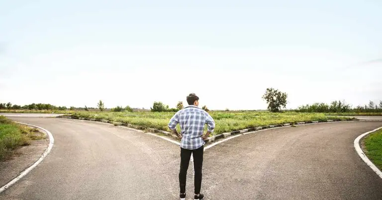 A man wearing blue jeans and a white and blue plaid collard shirt stands at the converging point of a large crossroads with large green expansive fields in the distance to symbolize career progression and whether to work contract of full-time.
