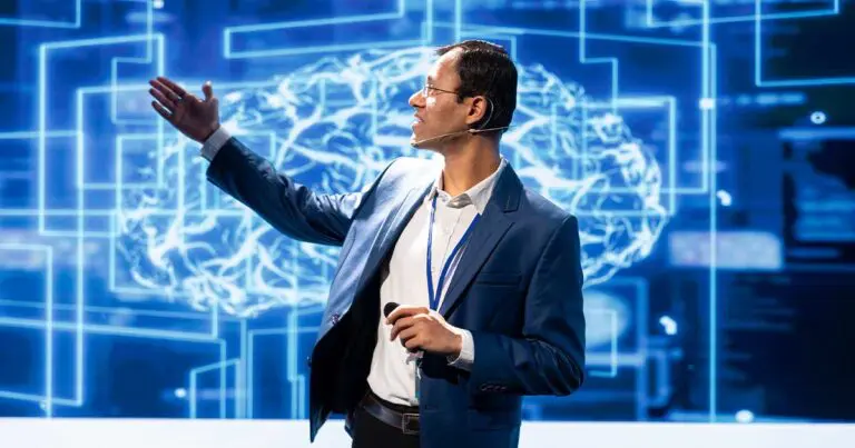 A man with dark hair and glasses wearing a blue blazer and a white collared shirt stands on a stage with blue digital images as his backdrop as he gestures to something out of frame as if he is giving a presentation to represent the future of cybersecurity hiring.