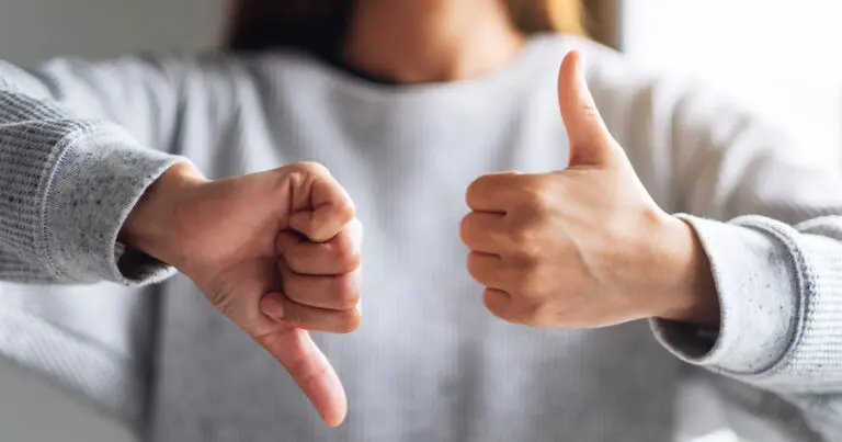 Image of a woman from the neck down with brown hair wearing a grey sweater in the background holding her hands up in front of her. The left is a thumbs down and the right is a thumbs up to symbolize how to ask for feedback at work.