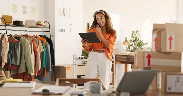 A young woman with brown curly hair wearing a flowing orange top and white pants happily talks on the phone while looking at a clipboard and surrounded by lots of shipment boxes in a bright naturally lit retail space to represent growing a small business