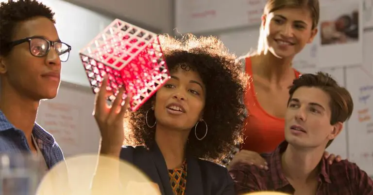 Two women and two men professionals sit around a table in a warm sunny room inspecting a gridded cube prototype to symbolize how to innovate in tech.