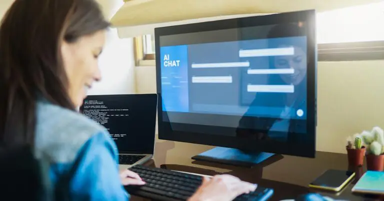 A woman with long brown hair wearing a denim blue shirt sits in front a computer with an AI chat interface on the screen while she smiles and types in her prompt to symbolize using ChatGPT to land a job.