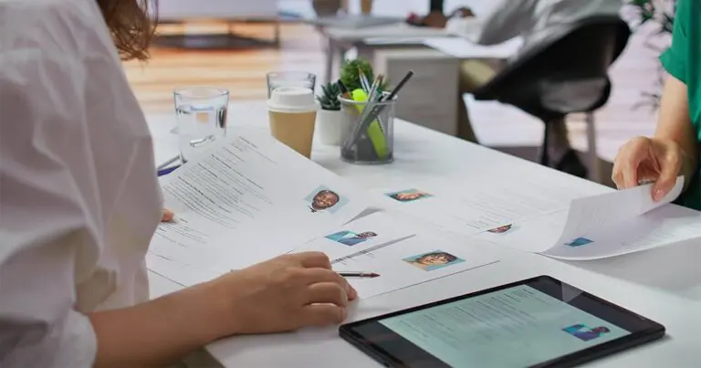 A professional in a white shirt sitting at a desk across another professional as they sort through resumes and job applications together to represent seasonal hiring trends