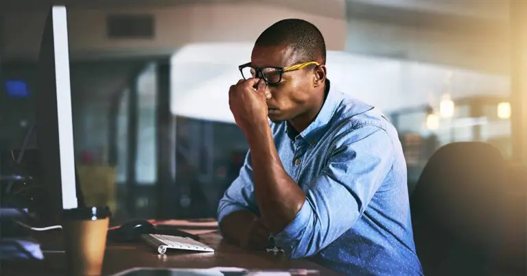 A professional man wearing a blue button up shirt sitting at a desk in front of a computer and pushing his glasses up to pinch the bridge of his nose as he breathes out in frustration to symbolize the pressures of being a VP or executive hiring under scrutiny