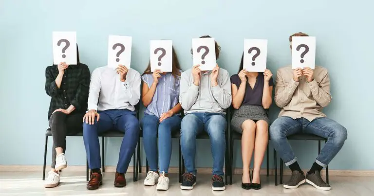 A row of job applicants sitting in chairs in a hallway, each holding a sign with a large question mark in front of their face to symbolize the unknowns of hiring a new candidate