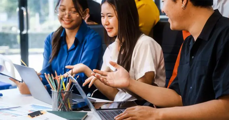 Two women and one male professional sit closely together at a small table in an office setting looking at their laptops together with a cup of colored pencils on the desk to symbolize how to innovate in tech when you're low on resources