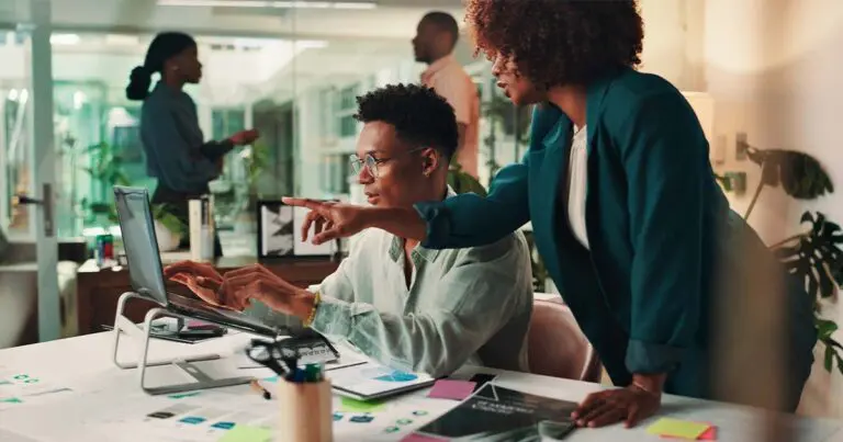 A young professional sits at a desk where a colleague stands next to him and points at his computer screen in a helpful manner to represent career building in your company