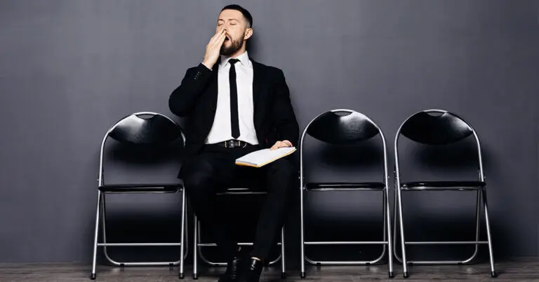 A professional in a black suit with a white shirt and tie sits yawning on a chair in a row of chairs in a hallway to represent candidate experience