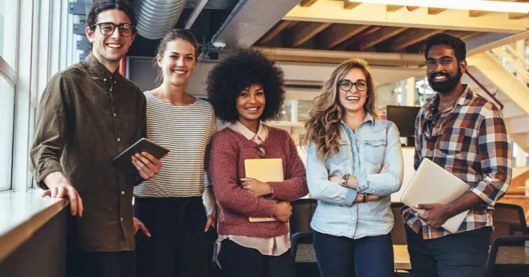 A diverse group of five smiling young professionals standing side-by-side in an industrial style office building dressed casually to signify a more relaxed work environment for AI implementation