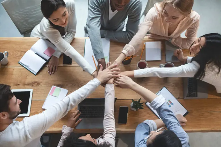 Top-down view of a diverse team stacking hands in unity around a table with laptops and notebooks, representing how to promote career building in your company through collaboration, trust, and inclusive culture.