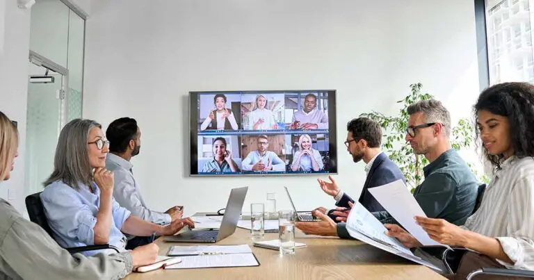Hybrid team of professionals in a modern conference room collaborating with remote colleagues on a video call—an example of how to promote career building in your company through flexible, inclusive, and cross-functional blended workforce models.