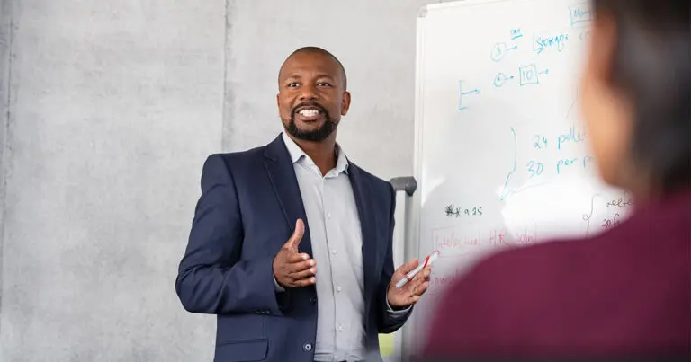 A professional business leader standing at the head of a room next to a white board and addressing to a group of people sitting blurred in the foreground to represent leadership training and upskilling