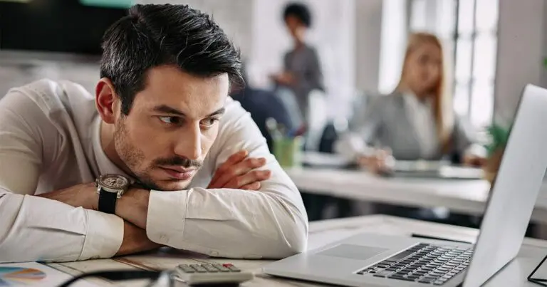 A man in a white dress shirt rests his chin on his arms, looking tired and unmotivated while staring at a laptop in an office setting. A woman in the background works at her desk, slightly out of focus.