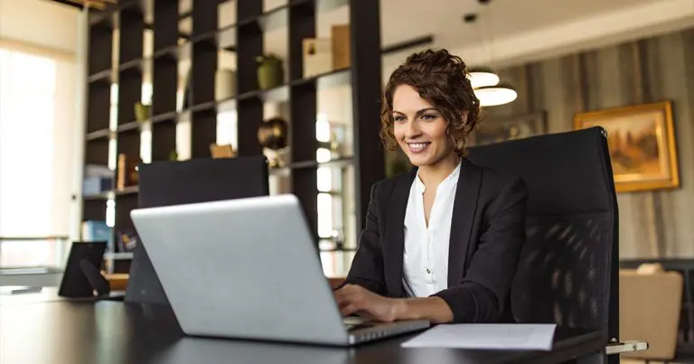 Confident professional woman smiling while working on a laptop in a modern office, representing how to promote career building in your company through flexible leadership models like fractional AI roles.