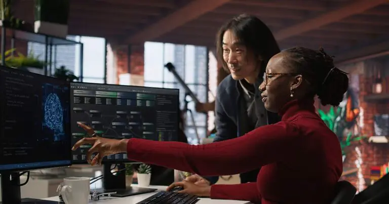 Two professionals analyzing AI system data on dual monitors, representing the value of fractional transformation leadership as a secret weapon for building AI-ready teams.
