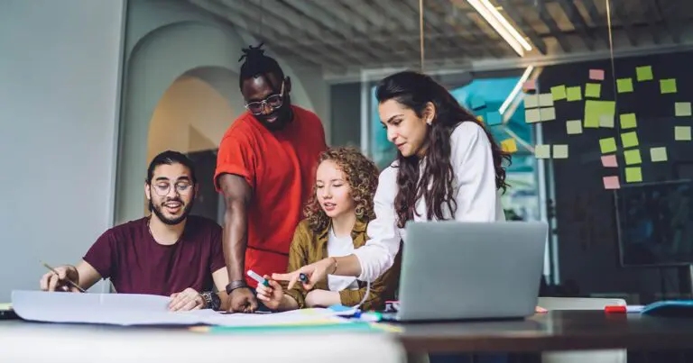 Four diverse young professionals collaborate around a large table in a modern office. One person points at a sheet of paper while others look on, engaged. A laptop, markers, and sticky notes are visible, with additional sticky notes covering the glass wall in the background.