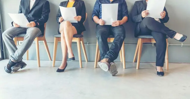 Four job candidates sit in a row of wooden chairs against a gray wall, holding printed resumes or documents in their laps. Only the lower half of their bodies is visible. They wear a mix of business and business-casual attire, including dress shoes, high heels, and sneakers. The setting suggests a waiting area for interviews, with neutral tones and minimal decor.