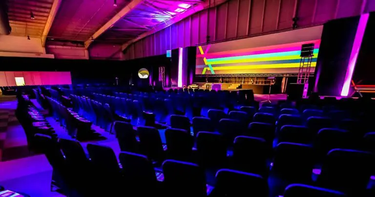 A large, empty conference hall with rows of black chairs facing a brightly lit stage. The stage features a wide screen with vivid horizontal stripes in pink, blue, yellow, and green. The lighting throughout the space is a dramatic mix of purple and pink, creating a vibrant atmosphere for a corporate or tech event.