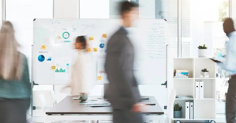 Modern office setting with several professionals walking past a large whiteboard covered in graphs, charts, sticky notes, and handwritten notes about sales and marketing strategy. The motion blur of the people walking conveys a fast-paced work environment. The room is bright and contemporary with natural light streaming through large windows.