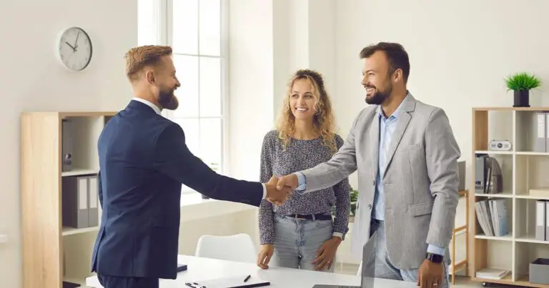 Two professionally dressed men shaking hands in an office with a smiling colleague nearby, symbolizing successful partnerships and how to promote career building in your company after consulting support ends.