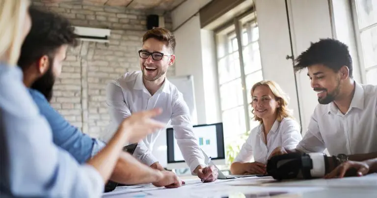 Group of five diverse professionals sitting around a conference table in a sunlit, modern office. They are smiling, engaged in discussion, and appear to be collaborating on a project. One man in glasses stands, leaning forward with enthusiasm as others listen and gesture.