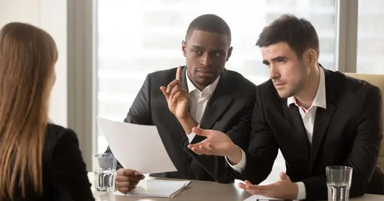 Two male interviewers in business suits sit at a table across from a female candidate. One man holds a resume and raises his index finger in concern while the other gestures skeptically, indicating disagreement or confusion. The candidate is seated with her back to the camera. The setting is a modern office with large windows.