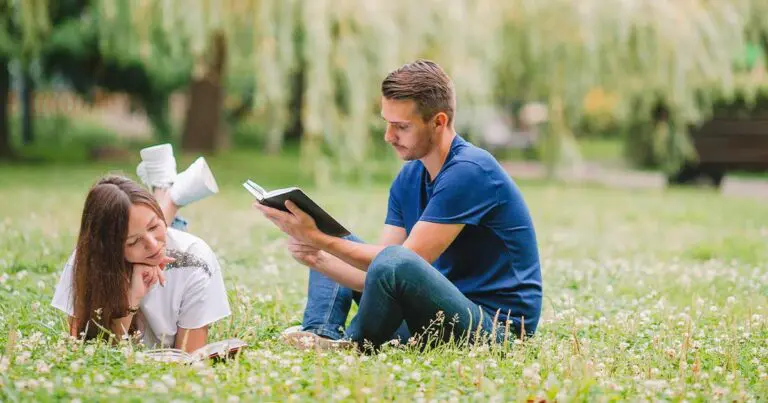 Two people sit on a grassy field reading books. A man in a navy t-shirt reads aloud from a book while a woman in a white t-shirt lies on her stomach next to him, listening and smiling. Trees and a park bench appear softly blurred in the background, suggesting a peaceful outdoor setting.