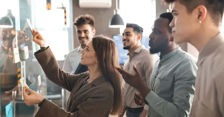 A diverse group of five young professionals collaborating in a modern office. A woman in a brown blazer places sticky notes on a glass wall, while her teammates—four men of varying ethnicities—look on and discuss ideas. The environment is casual and bright, with warm lighting and an atmosphere of teamwork and brainstorming.