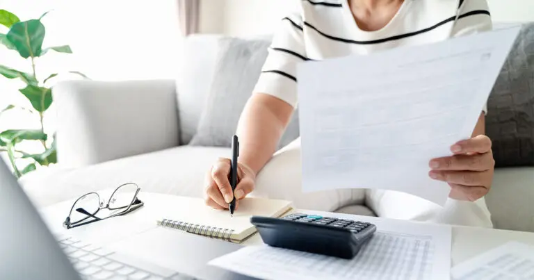 A person sitting on a couch reviews financial documents while taking notes on a notepad. A calculator, eyeglasses, and a laptop are visible on the coffee table in front of them, suggesting they are budgeting or working on financial planning from home.